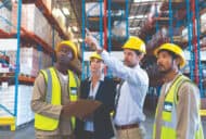 A group of people in a warehouse. They are wearing hardhats and safety vests.