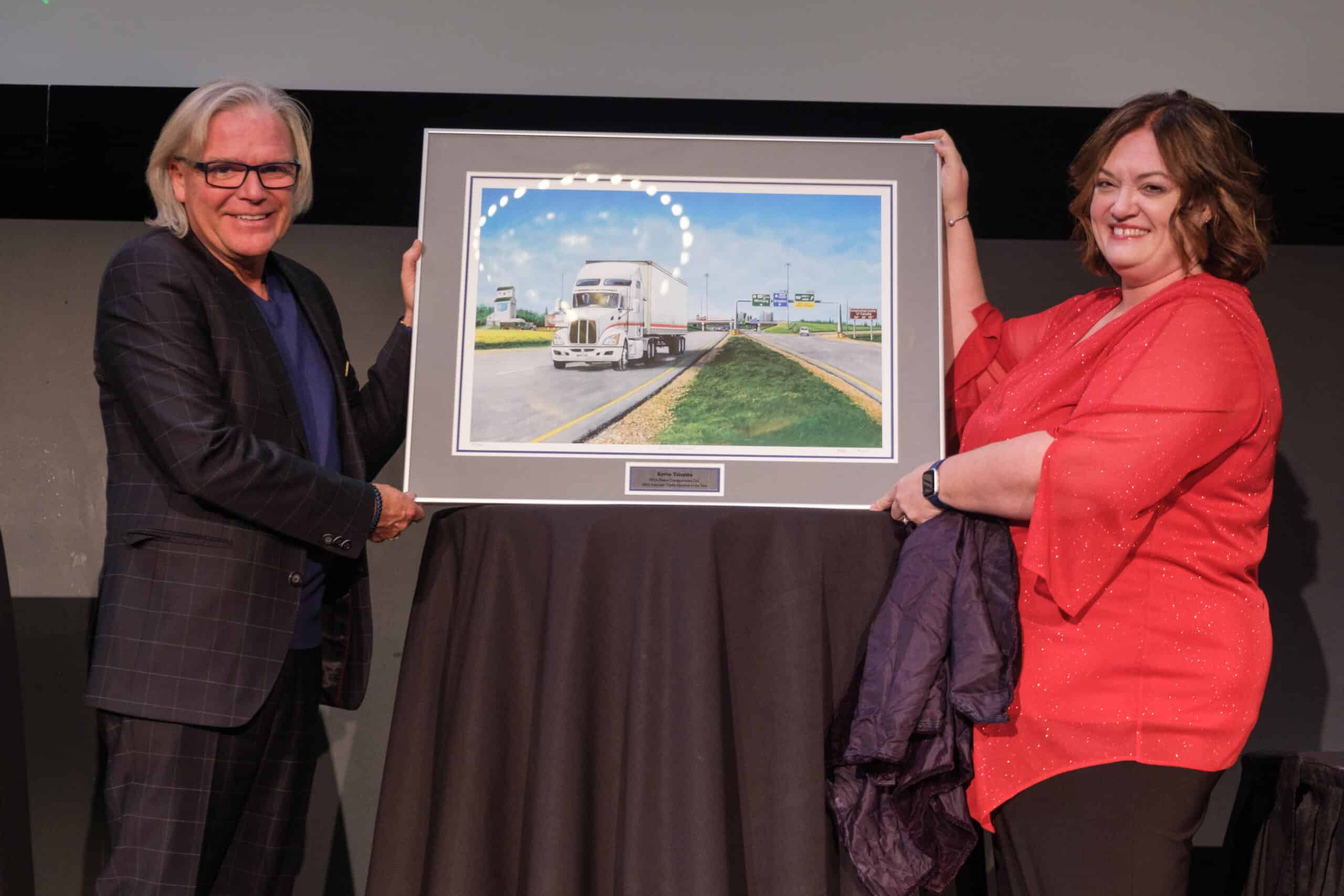Painting of a truck on a highway being presented on stage by Kevin Teixeira and Pauline Wiebe Peters