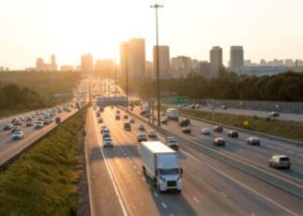 A picture of a white commercial truck and trailer travelling down a busy highway near a city.